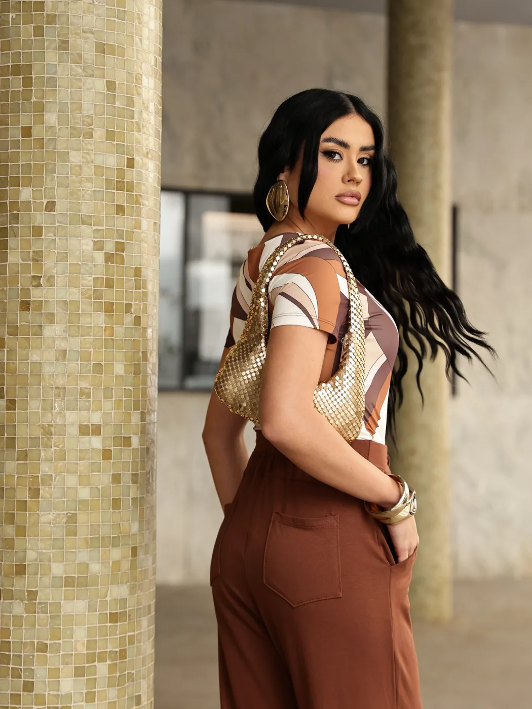 Raiany em look marrom ao lado de coluna de mosaico